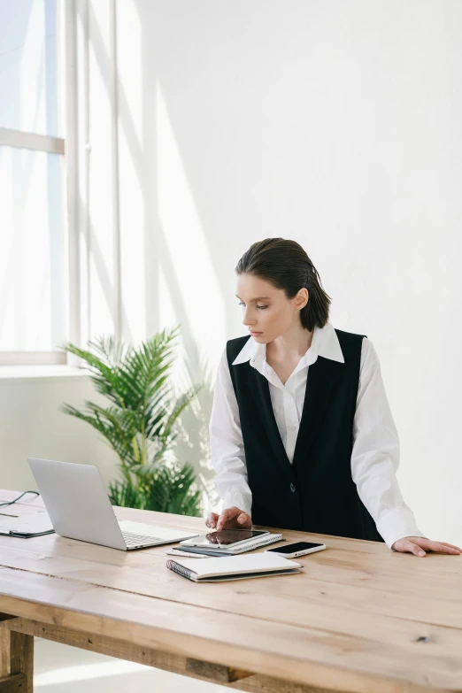a woman sitting at a table working on a laptop, wearing black vest and skirt, wearing a white button up shirt, in a white room, tailored clothing