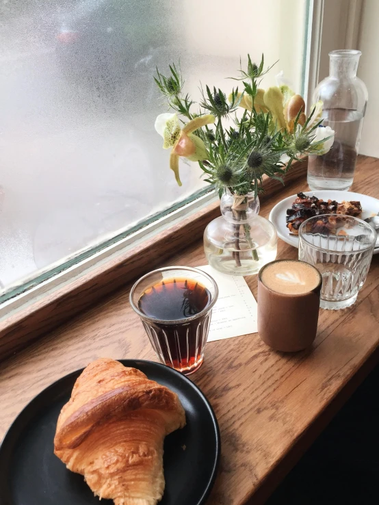 a black plate topped with a croissant next to a cup of coffee, photo of a beautiful window, cold brew coffee ), thumbnail, ignant