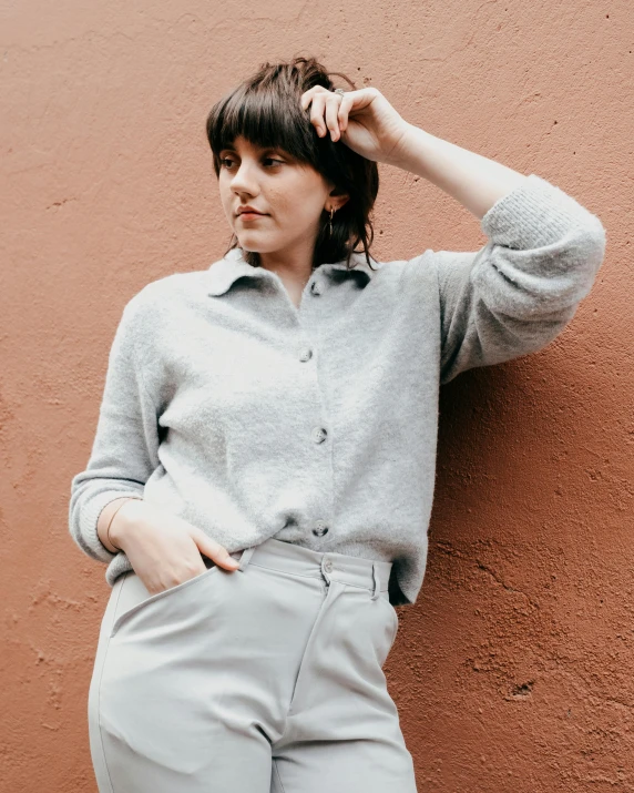 a woman leaning against a wall with her hand on her head, an album cover, by Helen Stevenson, trending on pexels, grey sweater, button up shirt, thumbnail, wearing pants