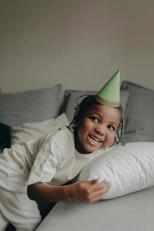 a little girl laying on a couch wearing a party hat, pexels contest winner, visual art, photo of a black woman, at a birthday party, green hat, wearing white pajamas