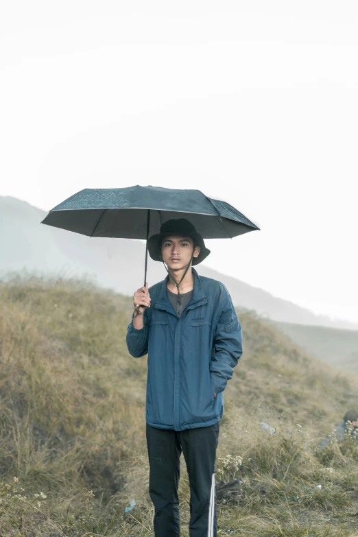 a man standing on a hill holding an umbrella, inspired by Zhang Kechun, unsplash, renaissance, wearing a navy blue utility cap, thin young male, avatar image, outdoor photo