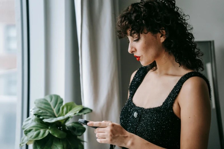 a woman looking at her cell phone by a window, by Julia Pishtar, trending on pexels, short black curly hair, wearing a sparkling dress, next to a plant, wearing black camisole outfit