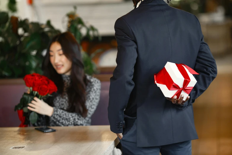 a man giving a gift to a woman at a table, pexels contest winner, white and red roses, backfacing, wearing a black and red suit, at checkout