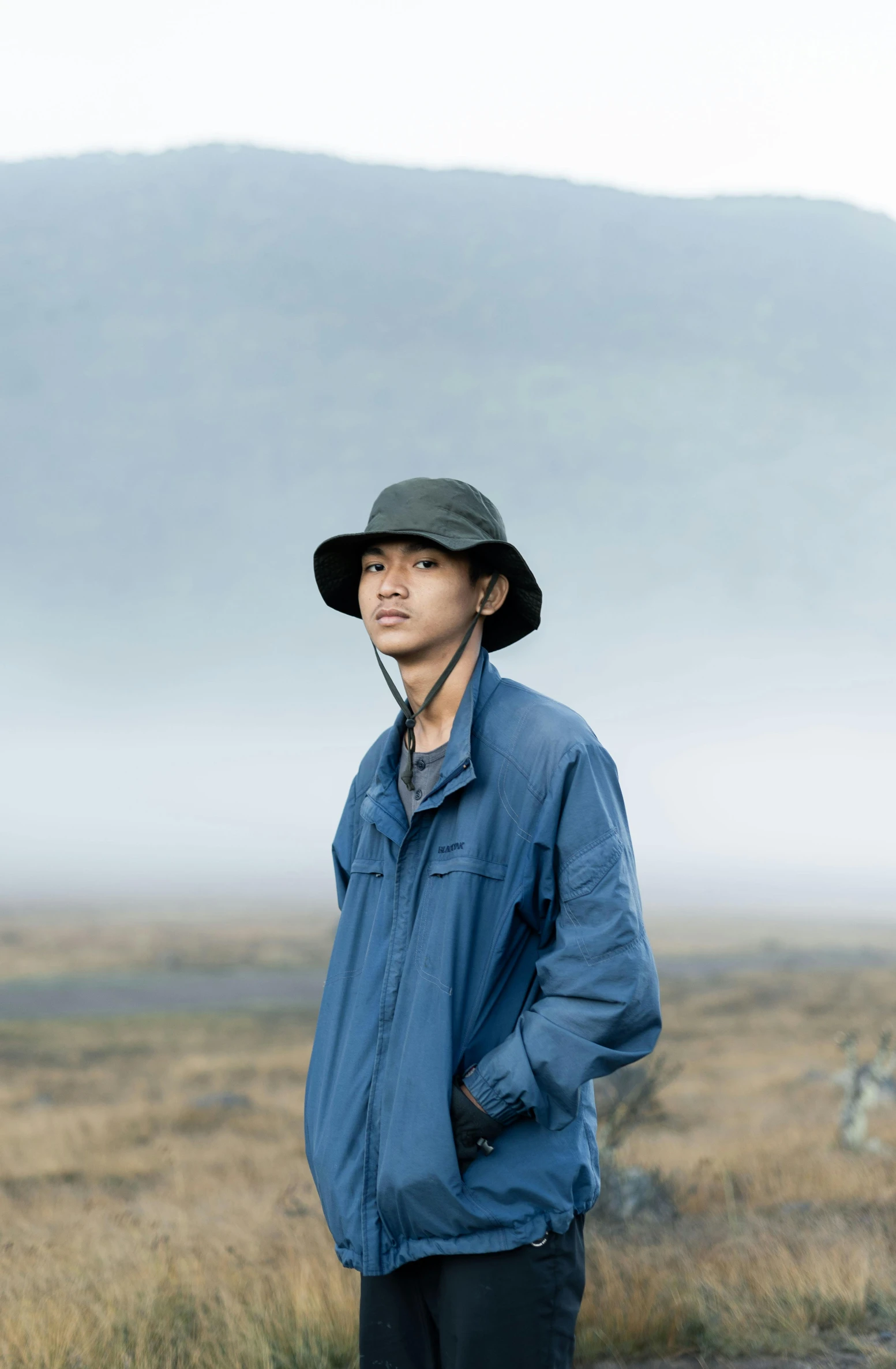 a man standing in the middle of a field, an album cover, inspired by Zhang Kechun, unsplash, realism, wearing a navy blue utility cap, iceland, portrait of young man, wearing a blue jacket