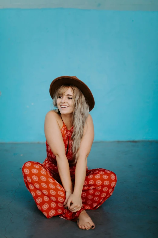 a woman sitting on the ground wearing a hat, trending on pexels, teal studio backdrop, red jumpsuit, concert photo, blonde