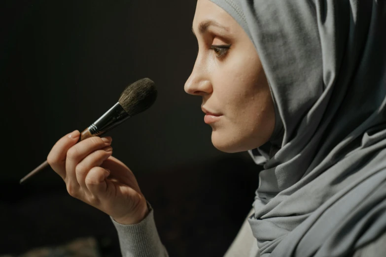 a woman in a hijab is using a makeup brush, pexels contest winner, hurufiyya, with grey skin, pale complexion, still from a movie, a woman's profile