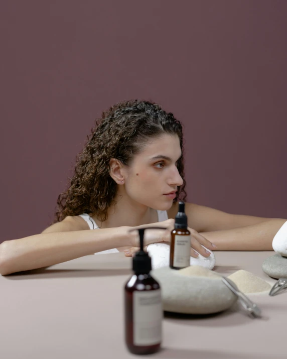 a woman sitting at a table with a bottle of lotion, trending on pexels, antipodeans, non binary model, looking serious, middle eastern skin, low quality photo