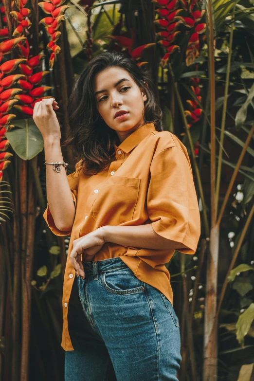 a woman standing in front of a tree with red flowers, a portrait, by Robbie Trevino, trending on pexels, wearing an orange t shirt, manila, wearing a linen shirt, caramel. rugged