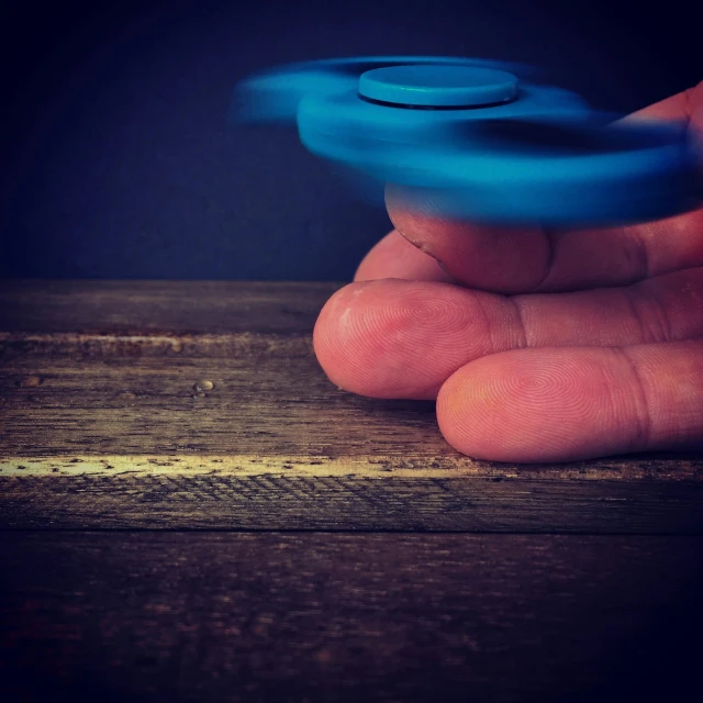 a hand holding a blue fidgetr on top of a wooden table, by Adam Marczyński, pexels contest winner, kinetic art, whirling, wheels, instagram picture, medium - shot