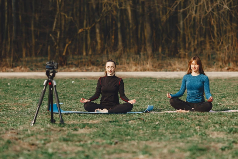 a couple of women sitting on top of a grass covered field, unsplash, video art, yoga, camera photo, against the backdrop of trees, avatar image