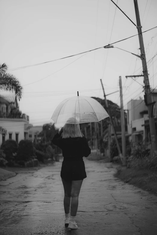 a woman walking down a street holding an umbrella, a black and white photo, facing away from camera, drippy, walking through a suburb, feeling miserable