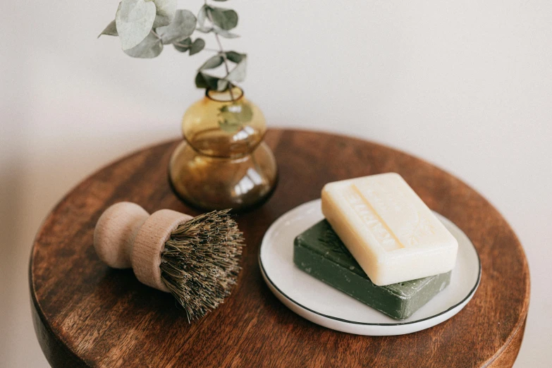 a couple of soap bars sitting on top of a wooden table, a still life, inspired by Géza Dósa, unsplash, plume made of seaweed, large brush, ceramic base, olive green