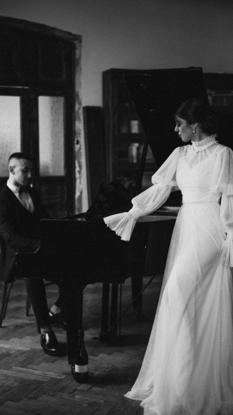 a black and white photo of a woman standing in front of a piano, a black and white photo, inspired by Hedda Sterne, unsplash, art nouveau, groom, wearing a white folkdrakt dress, live-action archival footage, alexander abdulov