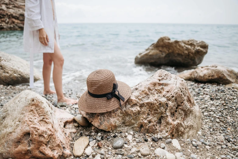 a woman standing on a rocky beach next to the ocean, pexels contest winner, renaissance, white straw flat brimmed hat, head down, brown, thumbnail