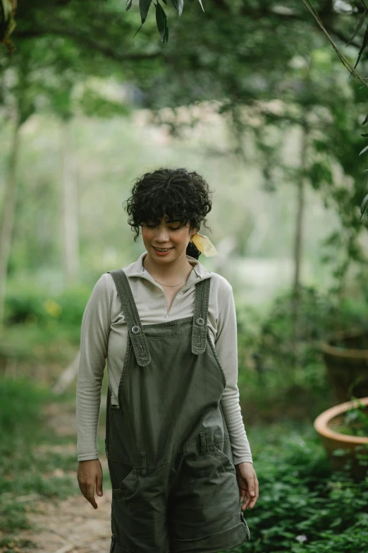 a woman in overalls walking down a path, inspired by Olive Mudie-Cooke, featured on instagram, renaissance, dark short curly hair smiling, in a verdant garden, still from film, blacksmith apron