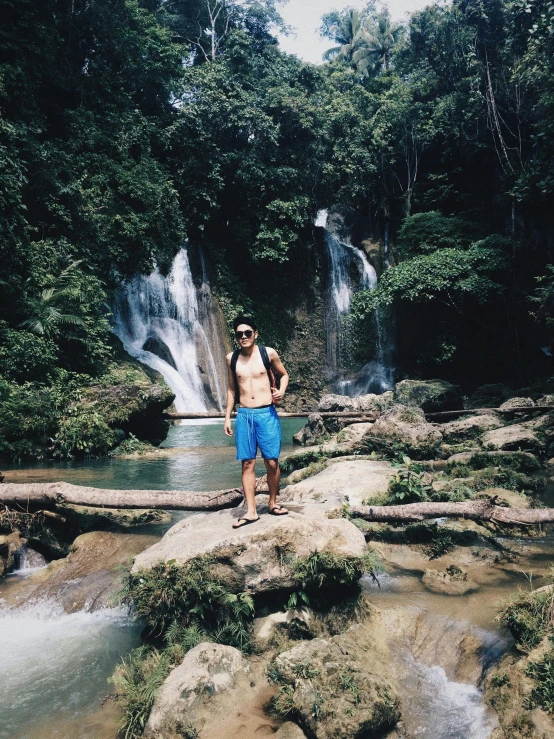 a man standing on a rock in front of a waterfall, inspired by Jorge Jacinto, sumatraism, 👰 🏇 ❌ 🍃, party in jungles, blue waters, with trees