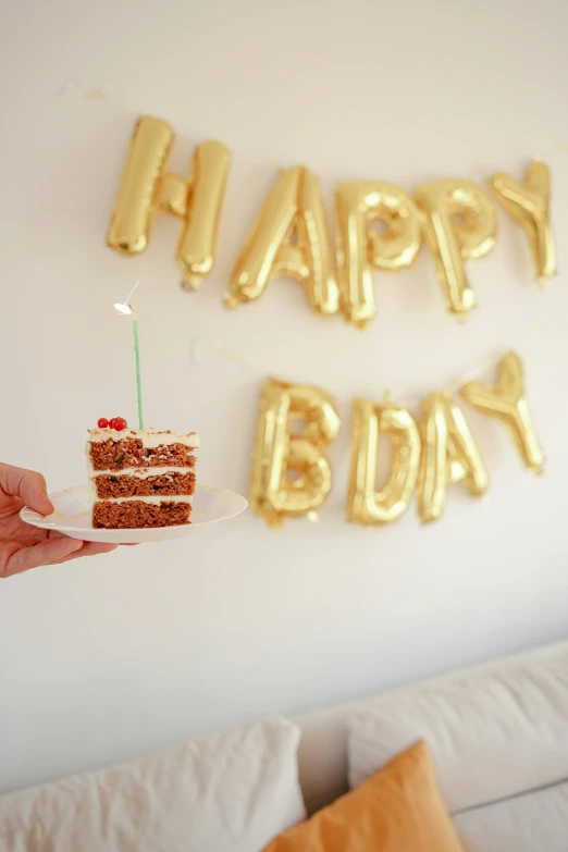 a person holding a piece of cake in front of a happy birthday banner, by Julia Pishtar, unsplash, shiny gold, balloon, in 2 0 1 5, dwell