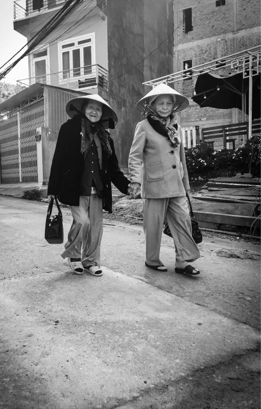 a black and white photo of two women walking down a street, by Yosa Buson, wearing a travel hat, hoang long ly, elderly, medium format