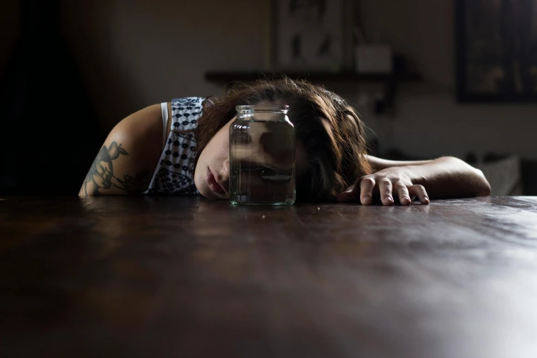 a woman laying on top of a wooden table next to a jar, pexels contest winner, tears running down, drink, julia sarda, laura watson