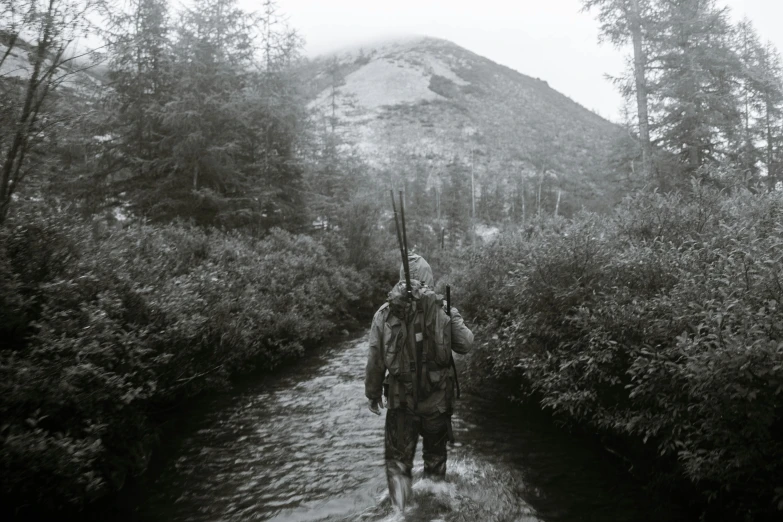 a black and white photo of a man walking across a river, hunters gear, sweaty mountain, dense thickets on each side, on the bow