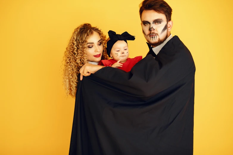 a man and woman in halloween costumes holding a baby, pexels, international gothic, yellow backdrop, vantablack cloak, square, blank