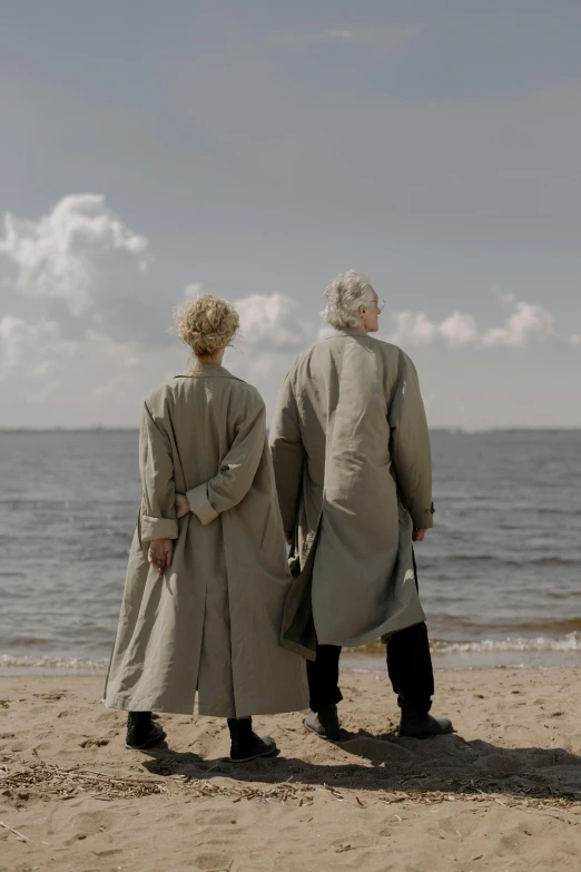 a couple of people standing on top of a sandy beach, an album cover, by Jan Tengnagel, unsplash, romanticism, trench coat, gray haired, ( ( theatrical ) ), bathrobe