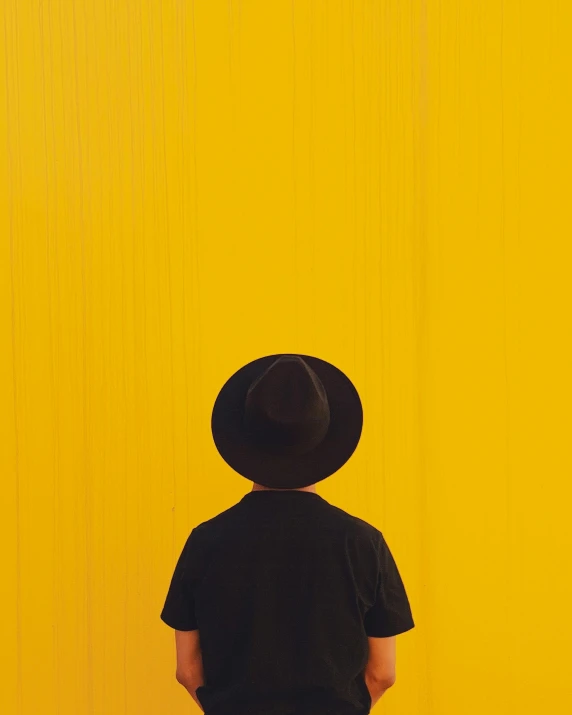 a person standing in front of a yellow wall, pexels contest winner, black hat, facing away, profile image, androgynous person