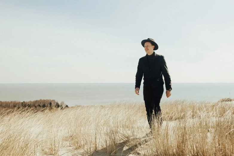 a man standing in a field next to the ocean, an album cover, unsplash, bauhaus, black stetson and coat, billy corgan, wearing black old dress and hat, prairie