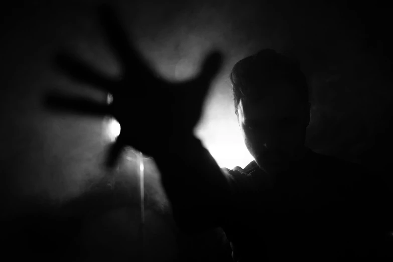 a person holding their hand up in the dark, a black and white photo, by Kristian Zahrtmann, pexels, bauhaus, backlit face, demon, album cover, silhouette of a man