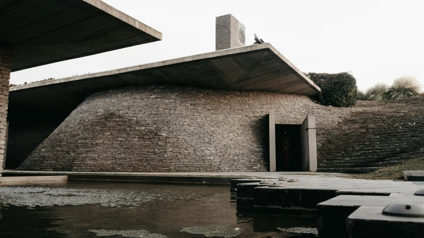 a building sitting next to a body of water, inspired by Peter Zumthor, unsplash, brutalism, stone roof, india, front side, government archive photograph