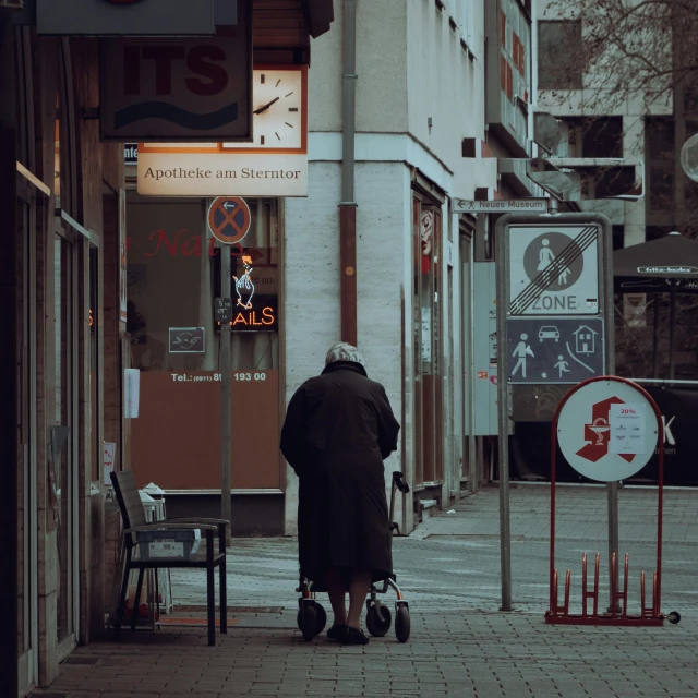 a person walking down a sidewalk with a stroller, by Tobias Stimmer, pexels contest winner, old lady, street signs, sorrow, wearing black overcoat