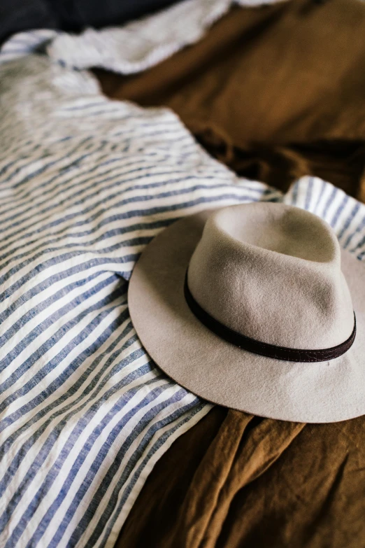 a hat sitting on top of a blanket on top of a bed, inspired by Joseph Beuys, unsplash, wearing a linen shirt, blue and grey theme, conde nast traveler photo, thumbnail