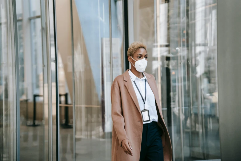 a woman wearing a face mask standing outside of a building, by Emma Andijewska, pexels contest winner, walking to work, african american woman, security agent, wearing a brown