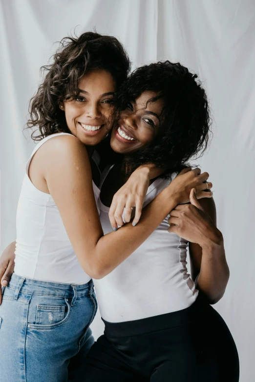 two women hugging each other in front of a white backdrop, trending on pexels, dark short curly hair smiling, roomies, gemini, black young woman