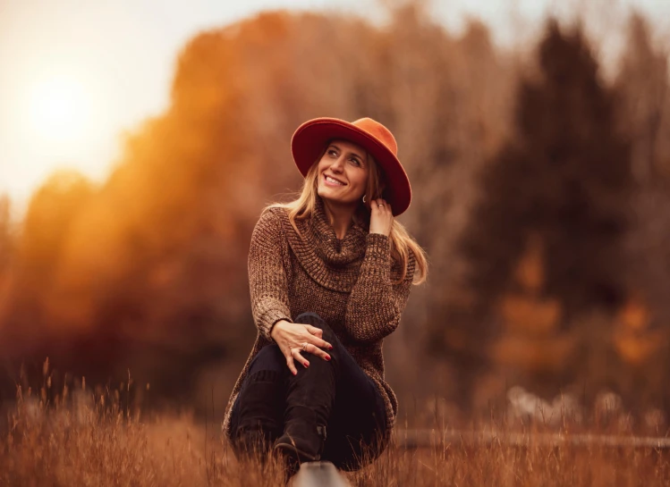 a woman in a hat is sitting in a field, pexels contest winner, warm color clothes, elegant smiling pose, autumn colours, beautifully backlit