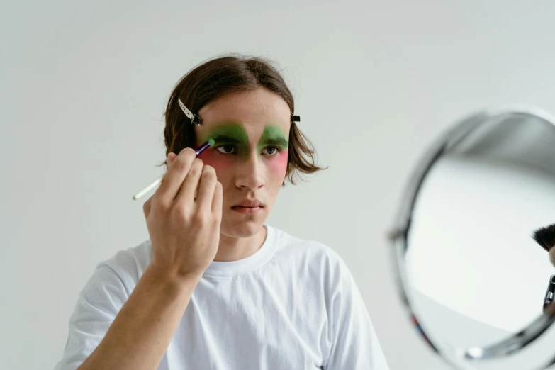 a woman putting makeup on her face in front of a mirror, inspired by Cindy Sherman, trending on pexels, man in a jester costume, skin painted with green, teenage boy, oval eyes