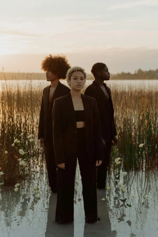 a group of people standing in front of a body of water, an album cover, by Winona Nelson, unsplash, renaissance, african american young woman, trio, in front of a forest background, dusk light