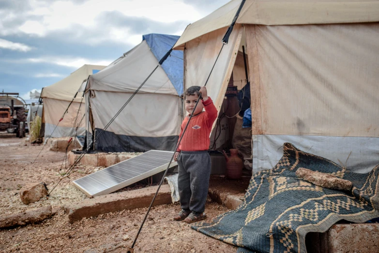 a man standing in front of a group of tents, hurufiyya, temporary emergency shelter, profile image, children, brown