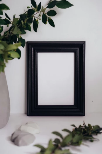 a picture frame sitting on top of a table next to a vase, by Julia Pishtar, all black matte product, on a white table, medium close up portrait, smooth panelling