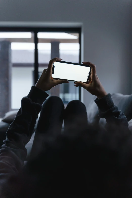 a person sitting on a couch holding up a cell phone, a picture, inspired by Elsa Bleda, trending on pexels, minimalism, crawling out of a dark room, front facing camera, slightly pixelated, spying