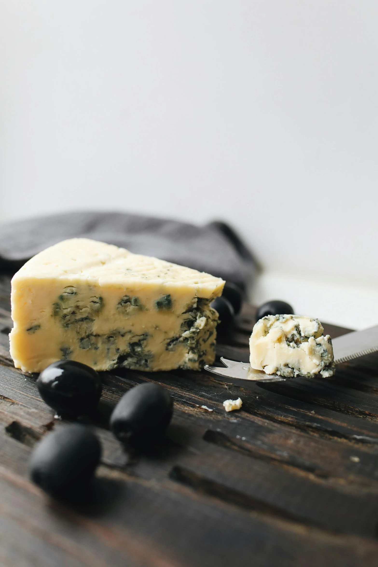 a piece of cheese sitting on top of a wooden cutting board, by Jessie Algie, renaissance, blue-black, olives, picton blue, umbra