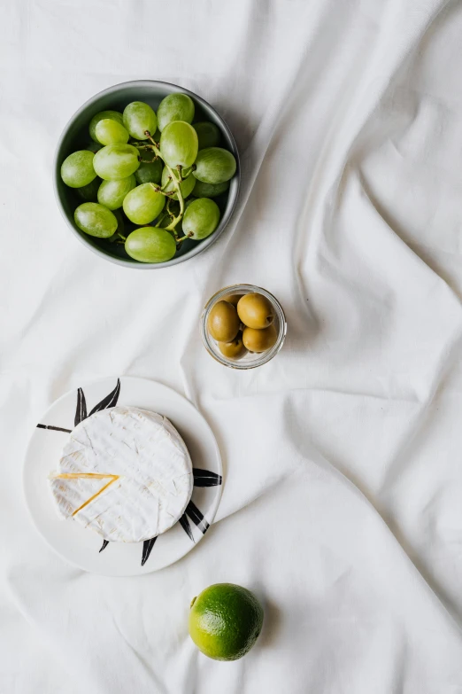 a white plate topped with grapes next to a bowl of fruit, inspired by Ceferí Olivé, unsplash, white bed, cheeses, patterned, white cloth