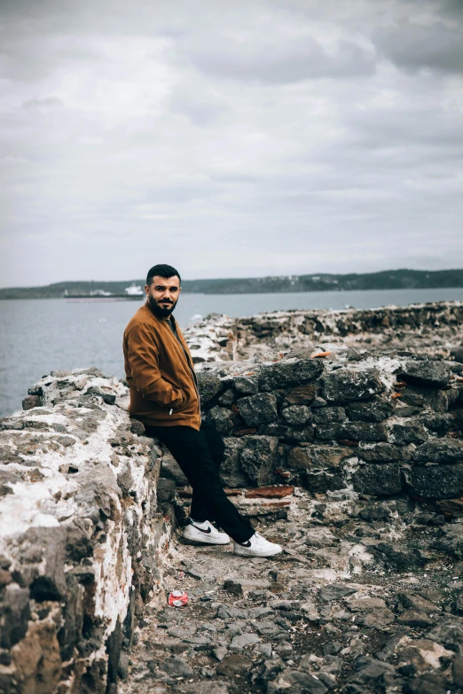a man sitting on a stone wall next to a body of water, an album cover, inspired by Oluf Høst, pexels contest winner, casual pose, a portrait of rahul kohli, in scotland, ocean view