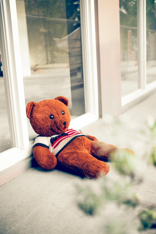 a brown teddy bear sitting in front of a window, by Julia Pishtar, patriotism, with a hurt expression, leaning on door, grieving