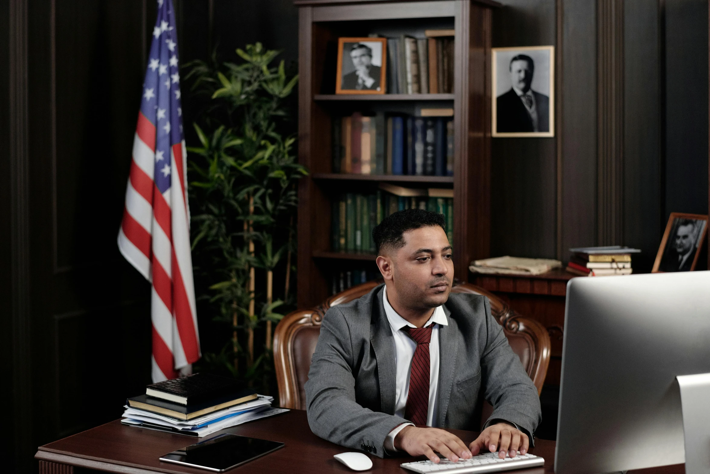 a man sitting at a desk in front of a computer, american flag in background, lawyer clothing, ismail, high quality photo