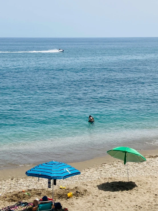 a couple of umbrellas sitting on top of a sandy beach, people swimming, malibu canyon, crystal clear blue water, small boat in foreground