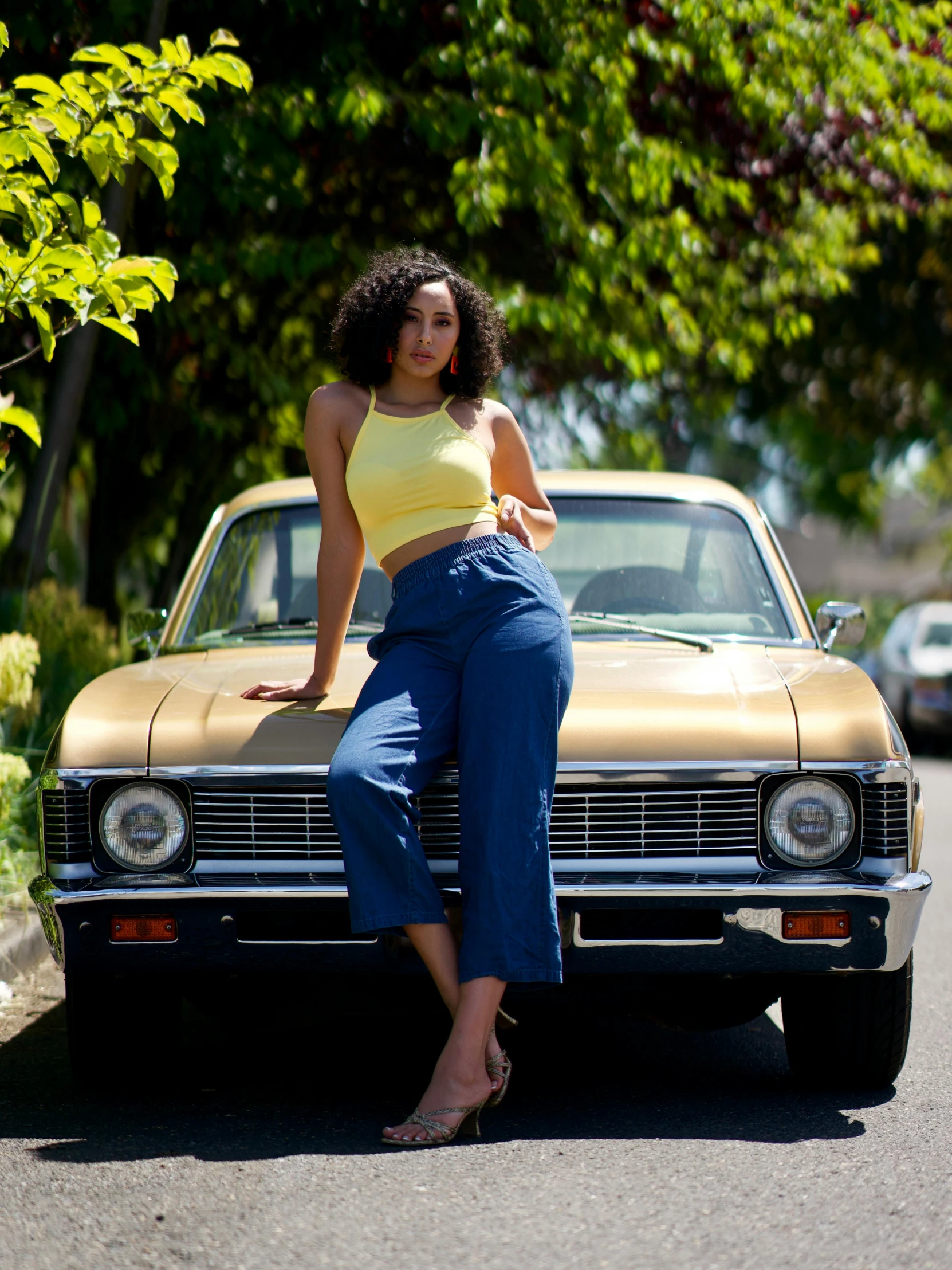 a woman sitting on the hood of a car, by Gwen Barnard, pexels contest winner, golden blue outfit, blue pants, wearing yellow croptop, female camila mendes