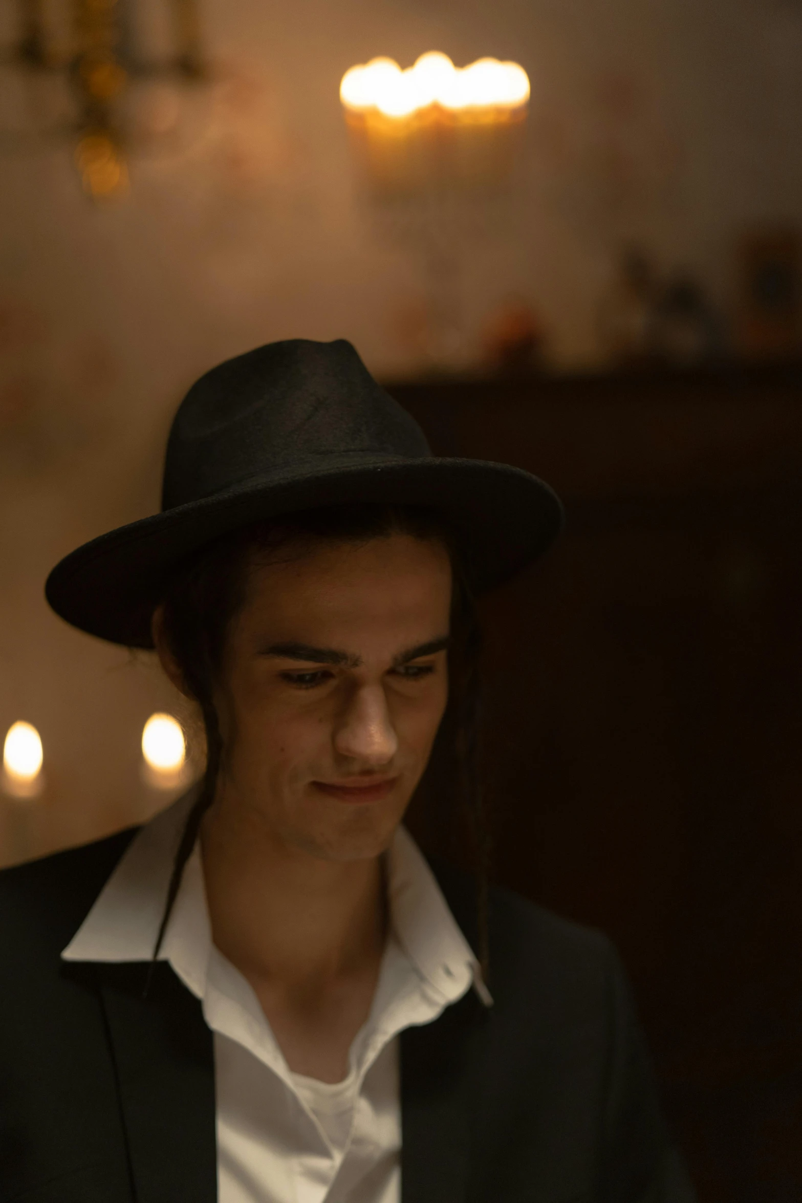 a man wearing a black hat and a white shirt, inspired by Nan Goldin, unsplash, antipodeans, avan jogia angel, in a room full of candles, ( ( theatrical ) ), wearing a fedora