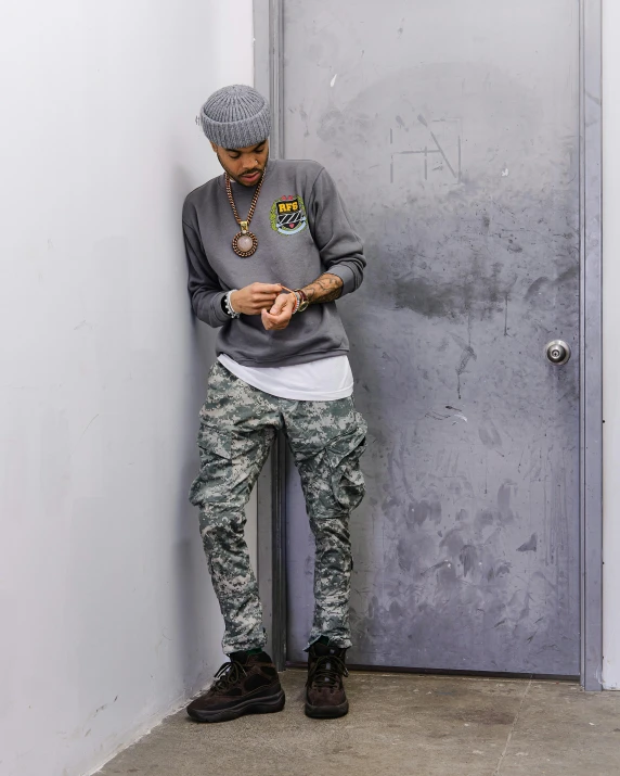 a man leaning against a wall looking at his cell phone, inspired by Carl Gustaf Pilo, graffiti, multicam uniform, grey sweater, wiz khalifa, standing on 2 feet