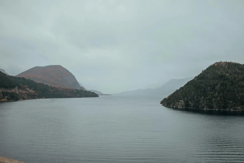 a body of water with mountains in the background, grey sky, during autumn, fjords, 8 k film still